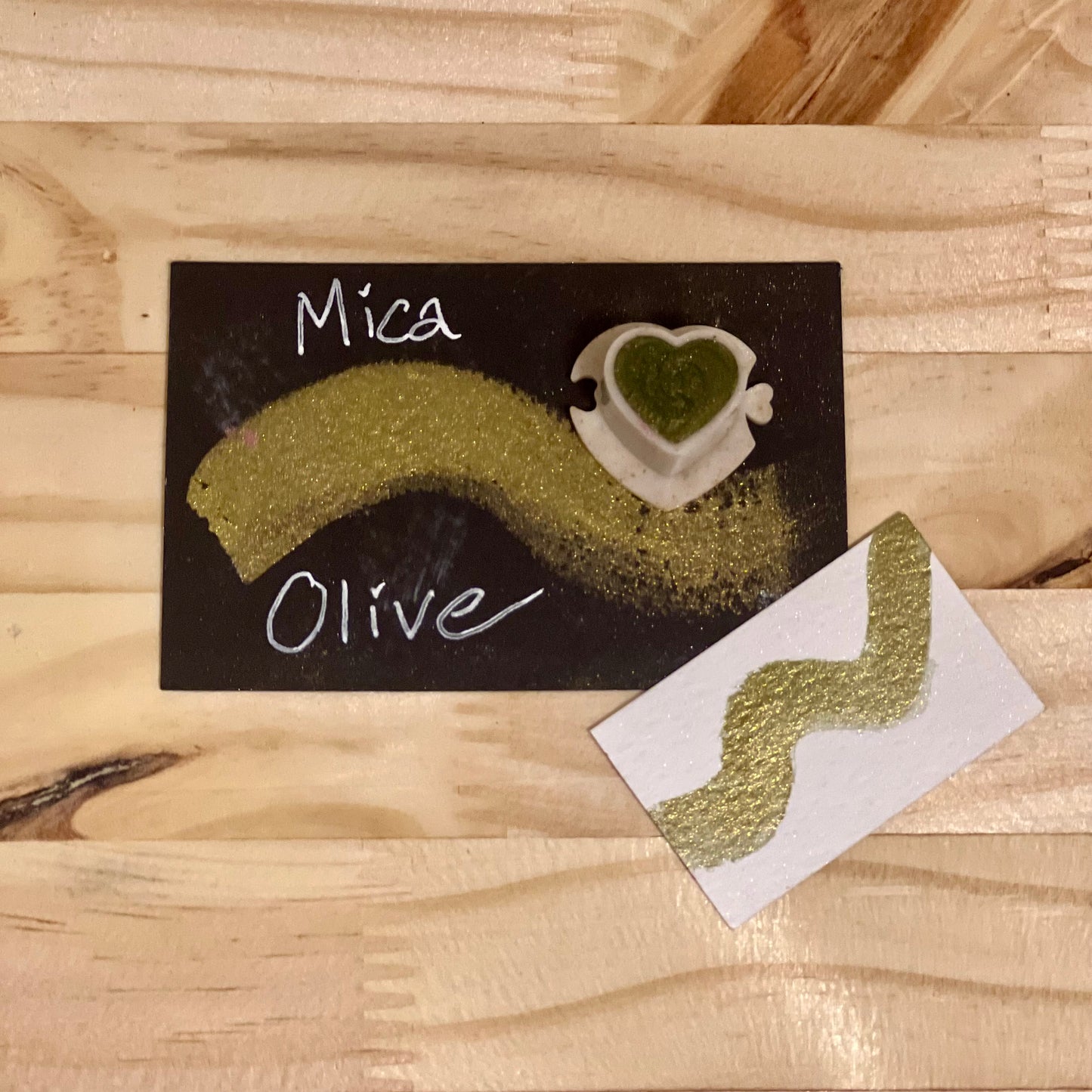 Olive mica shimmer watercolor paint heart shape paint pan with swatch on on a black card with 'Mica Olive' text and small swatch on white paper
