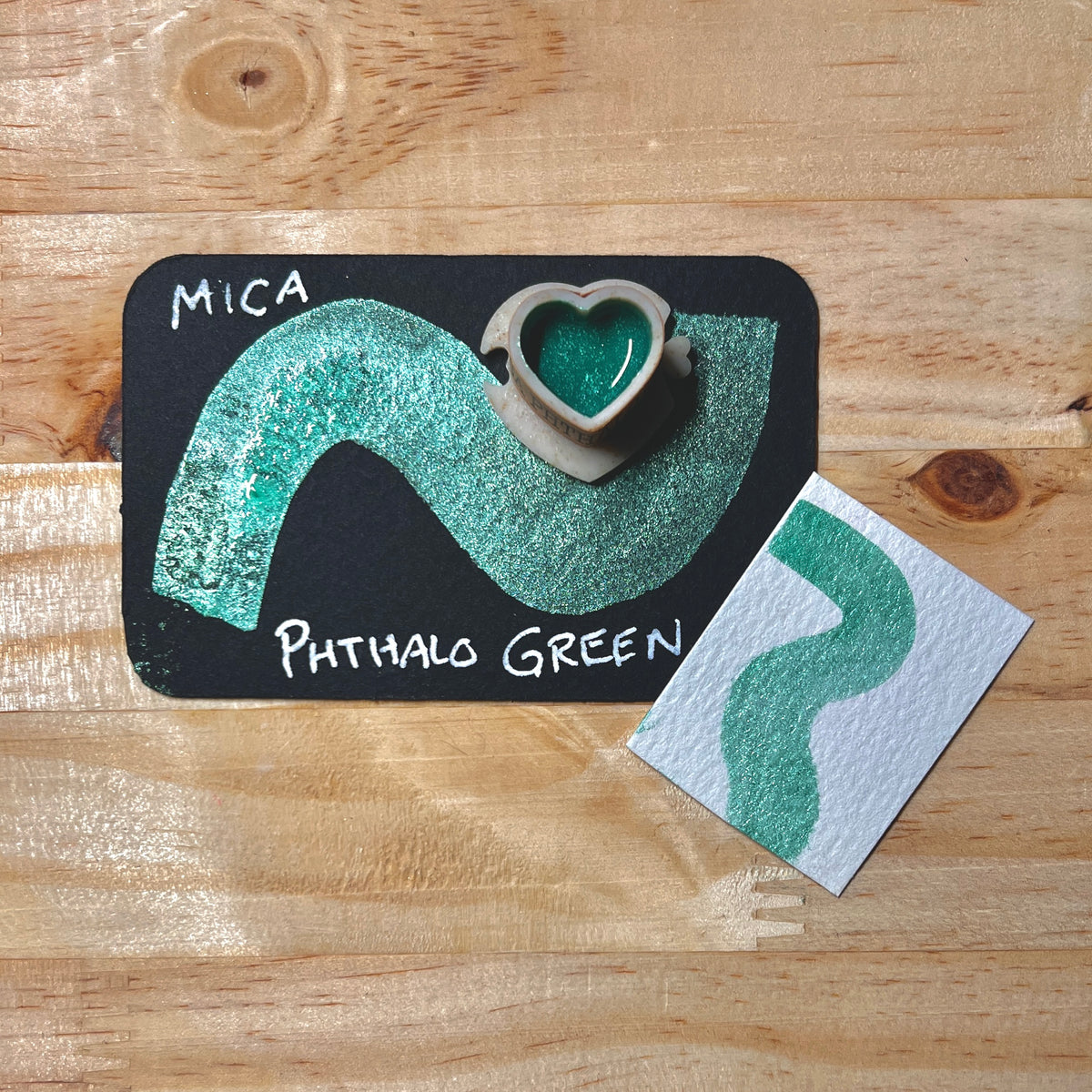 Phthalo green mica shimmer watercolor paint heart shape paint pan with swatch on on a black card with 'Mica Phthalo Green' text and small swatch on white paper