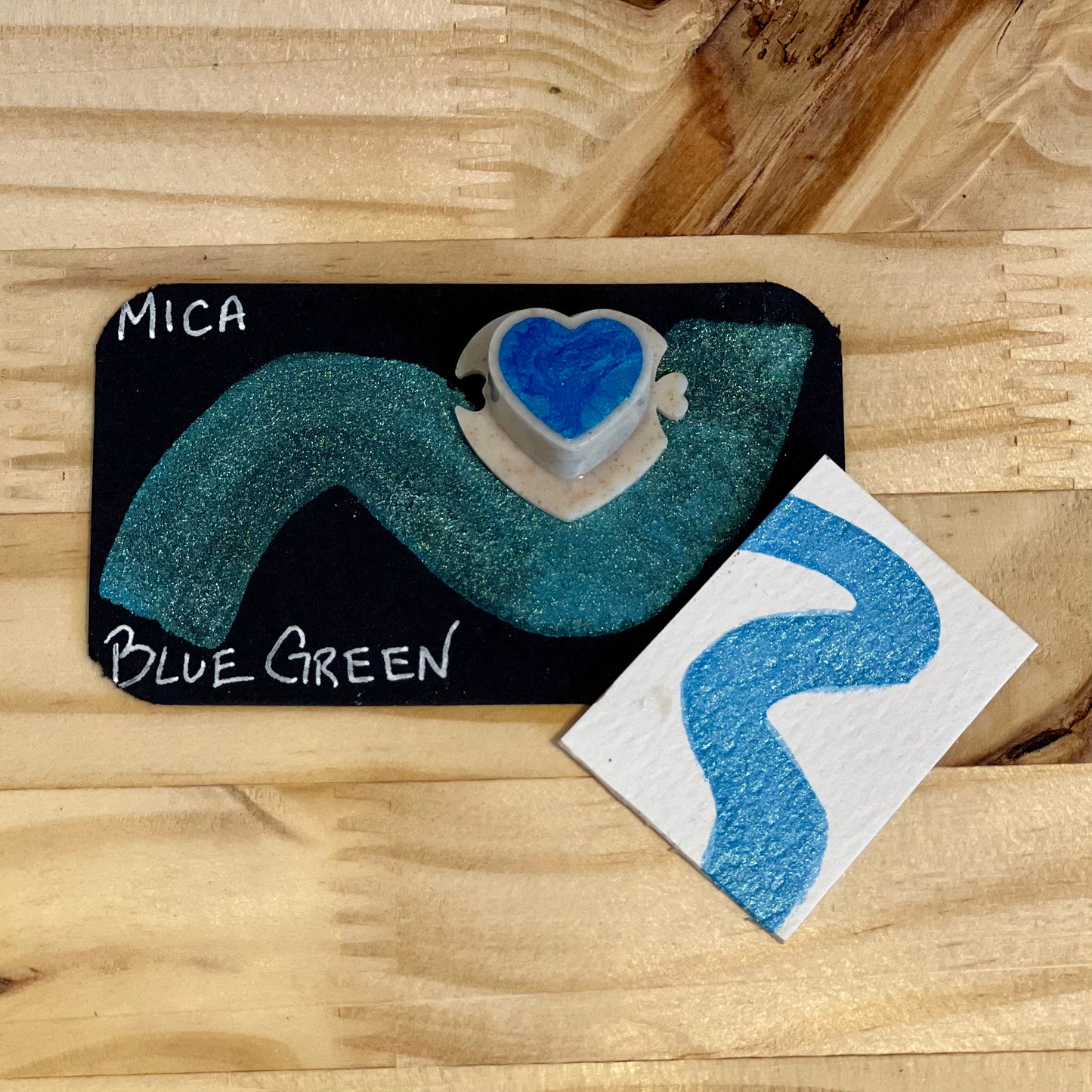 blue green mica shimmer watercolor paint  heart shape paint pan with swatch on on a black card with 'Mica Blue Green' text and small swatch on white paper
