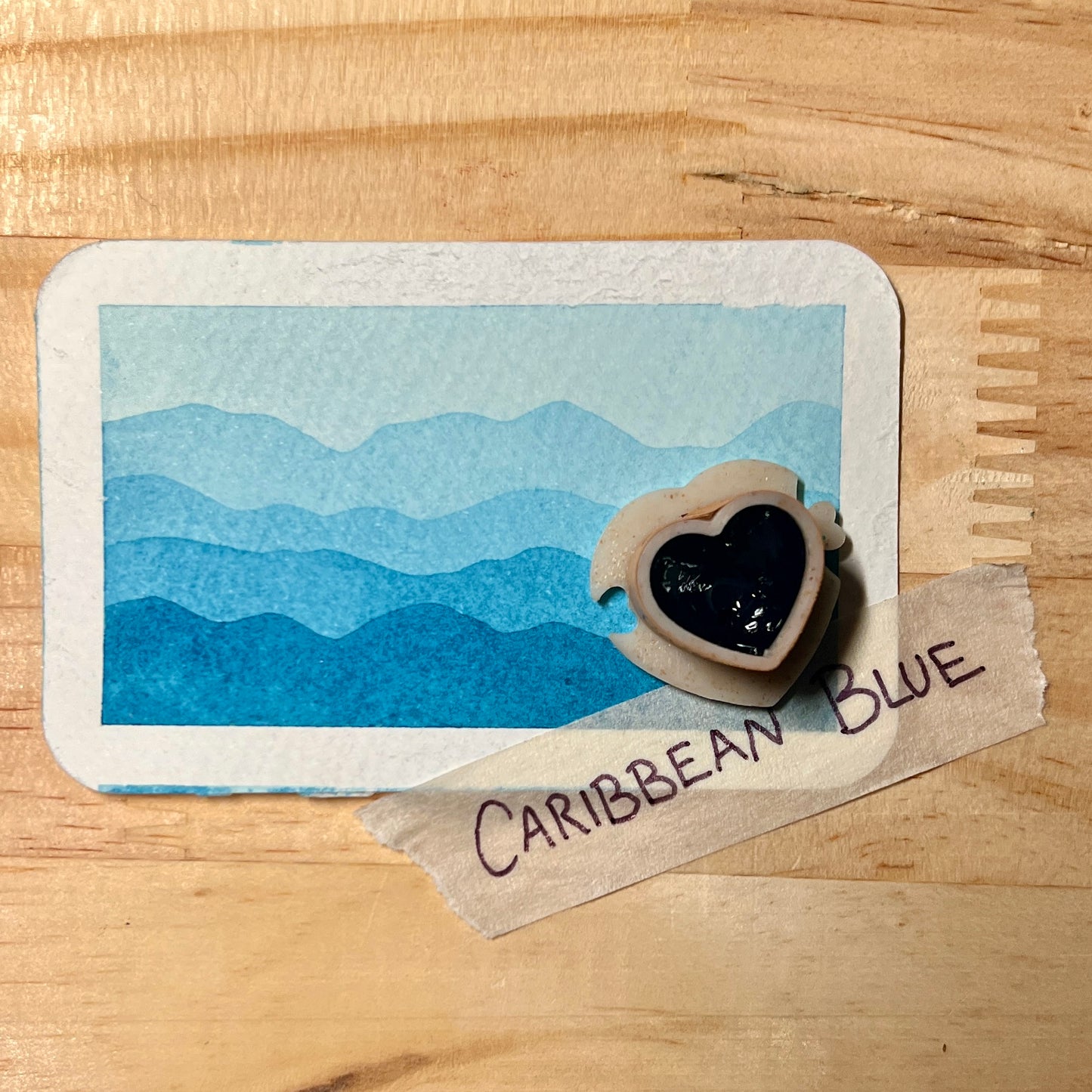 Card with blue mountain design and heart-shaped object on a wooden surface, labeled 'Caribbean Blue'.
