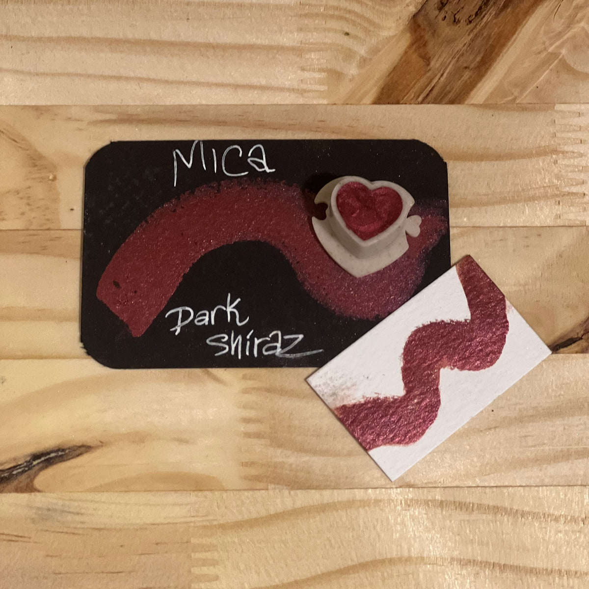 Dark Shira mica shimmer watercolor paint  heart shape paint pan with swatch on on a black card with 'Mica Dark Shiraz' text and small swatch on white paper