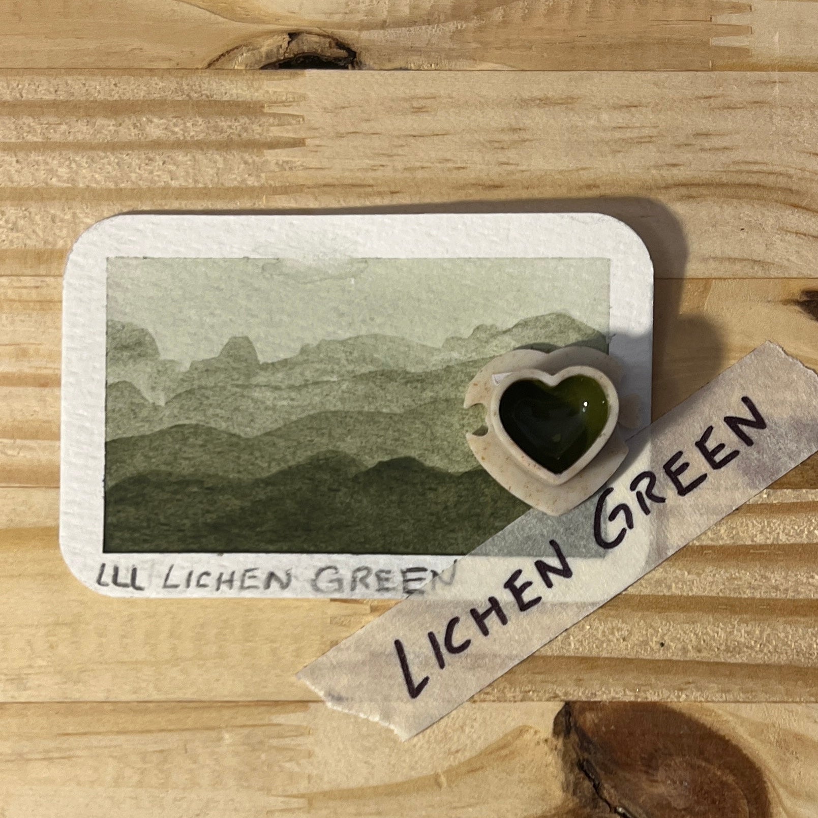 Card with landscape illustration and heart-shaped pan on a wooden surface with 'Lichen Green' label text.