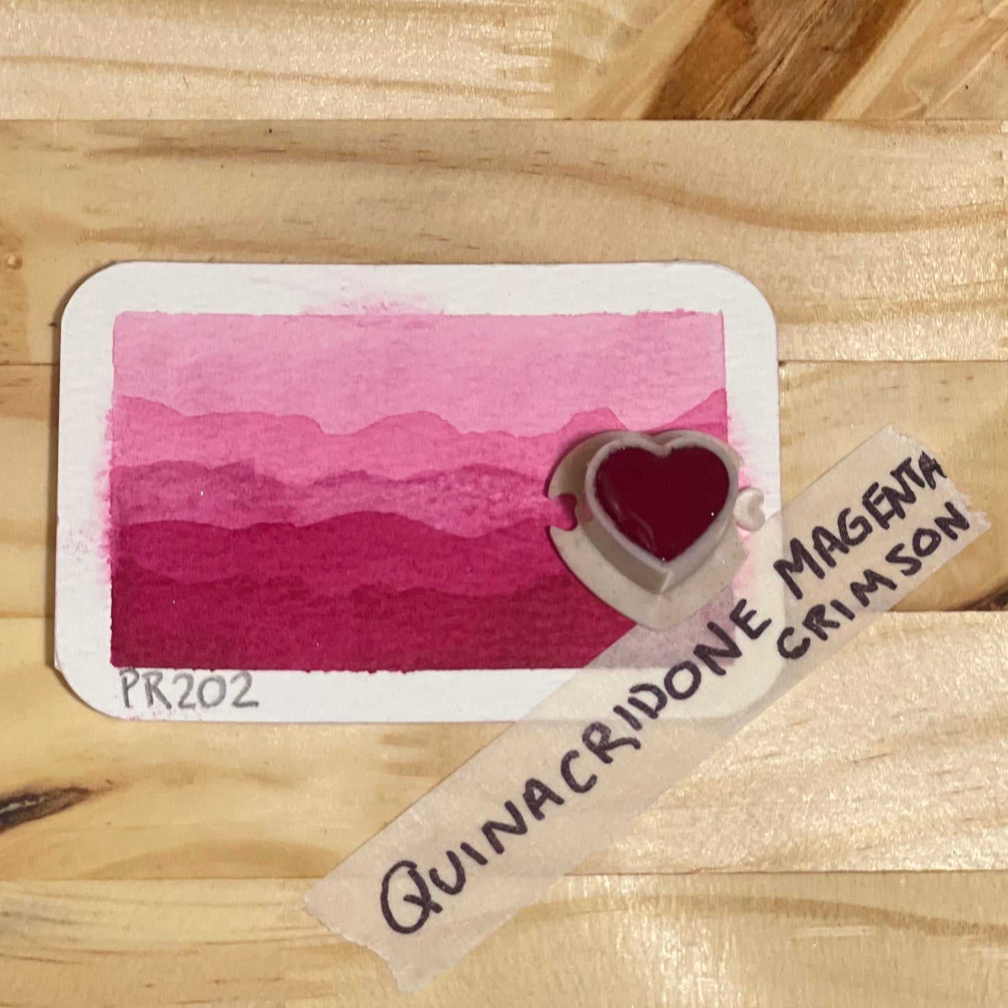 magenta crimson watercolor gradient landscape, heart-shape watercolor pan, labeled 'Quinacridone Magenta Crimson', on a wooden surface.