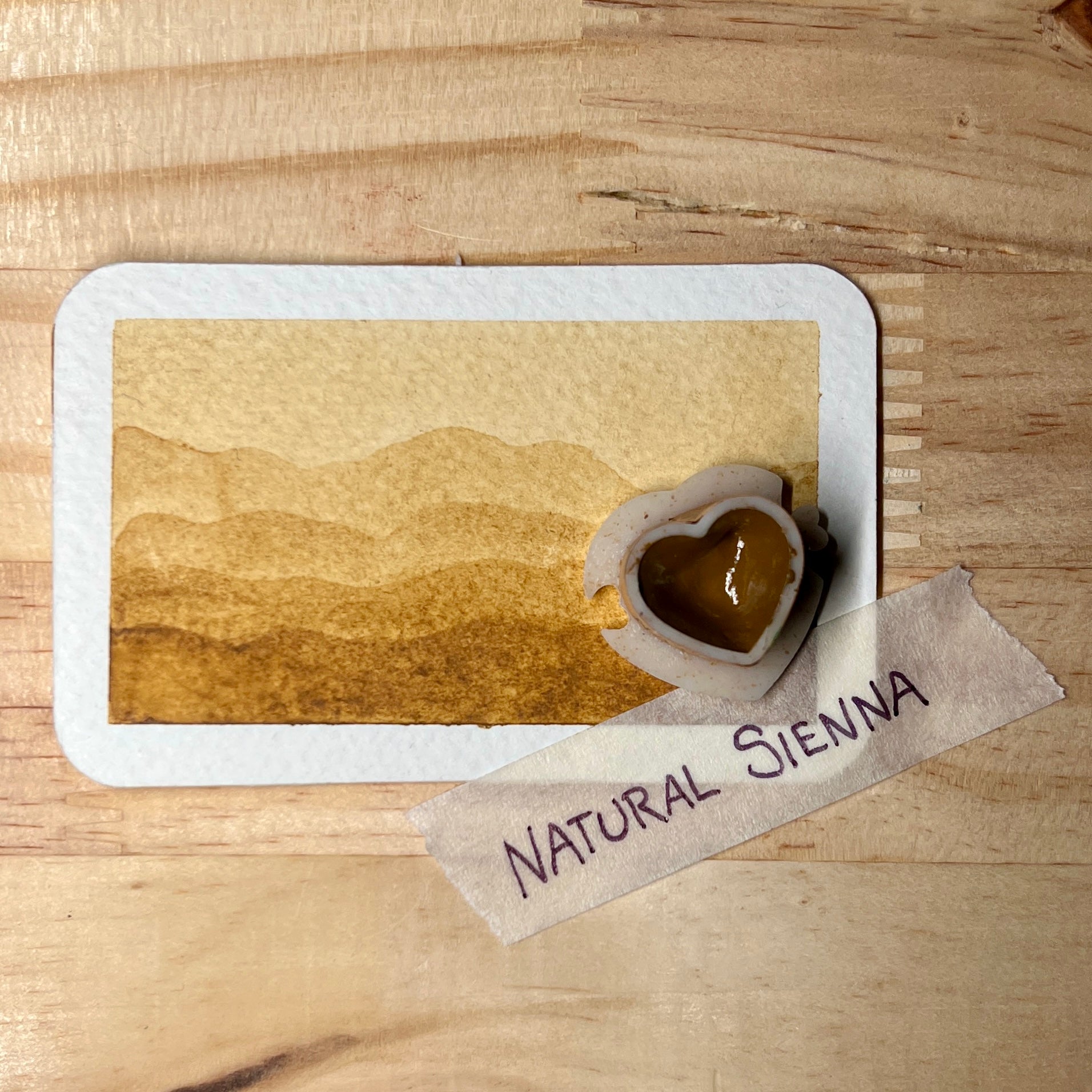 Card with a heart-shaped watercolor pan and watercolor landscape on a wooden surface, labeled 'Natural Sienna'.