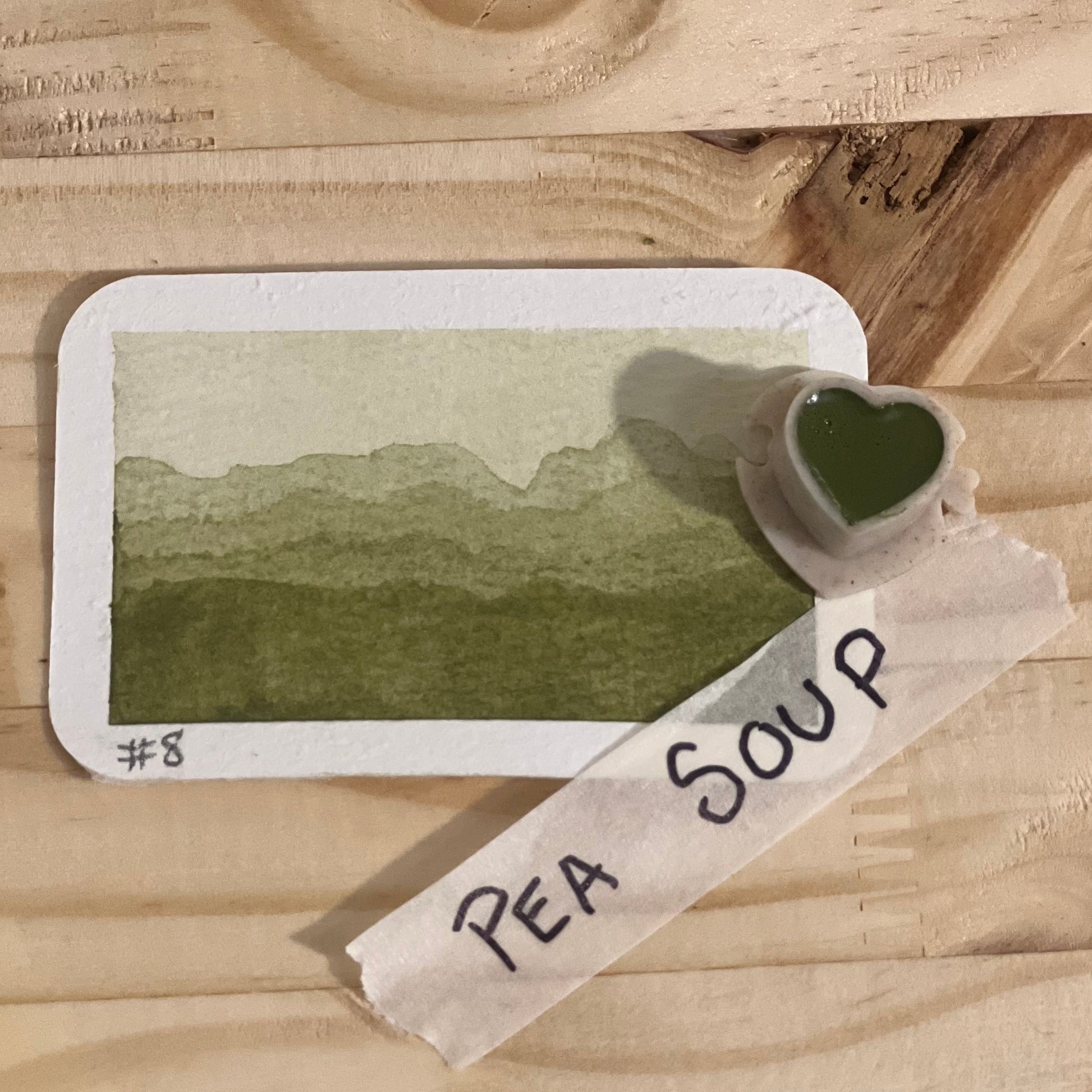 olive green watercolor gradient landscape, heart-shape watercolor pan, labeled 'Pea soup', on a wooden surface.
