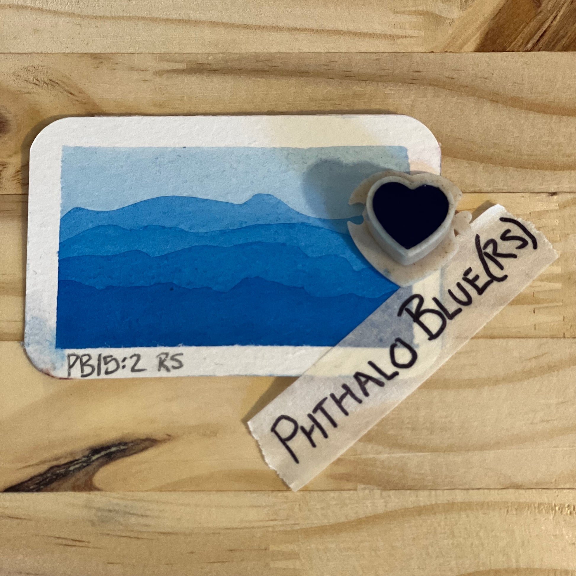 blue watercolor gradient landscape, heart-shape watercolor pan, labeled 'Phthalo Blue (RS)', on a wooden surface.