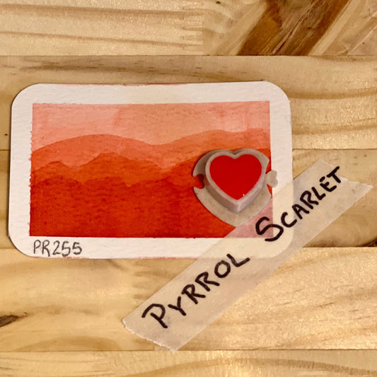 Color swatch labeled 'Pyrrol Scarlet' with a heart symbol on a wooden surface