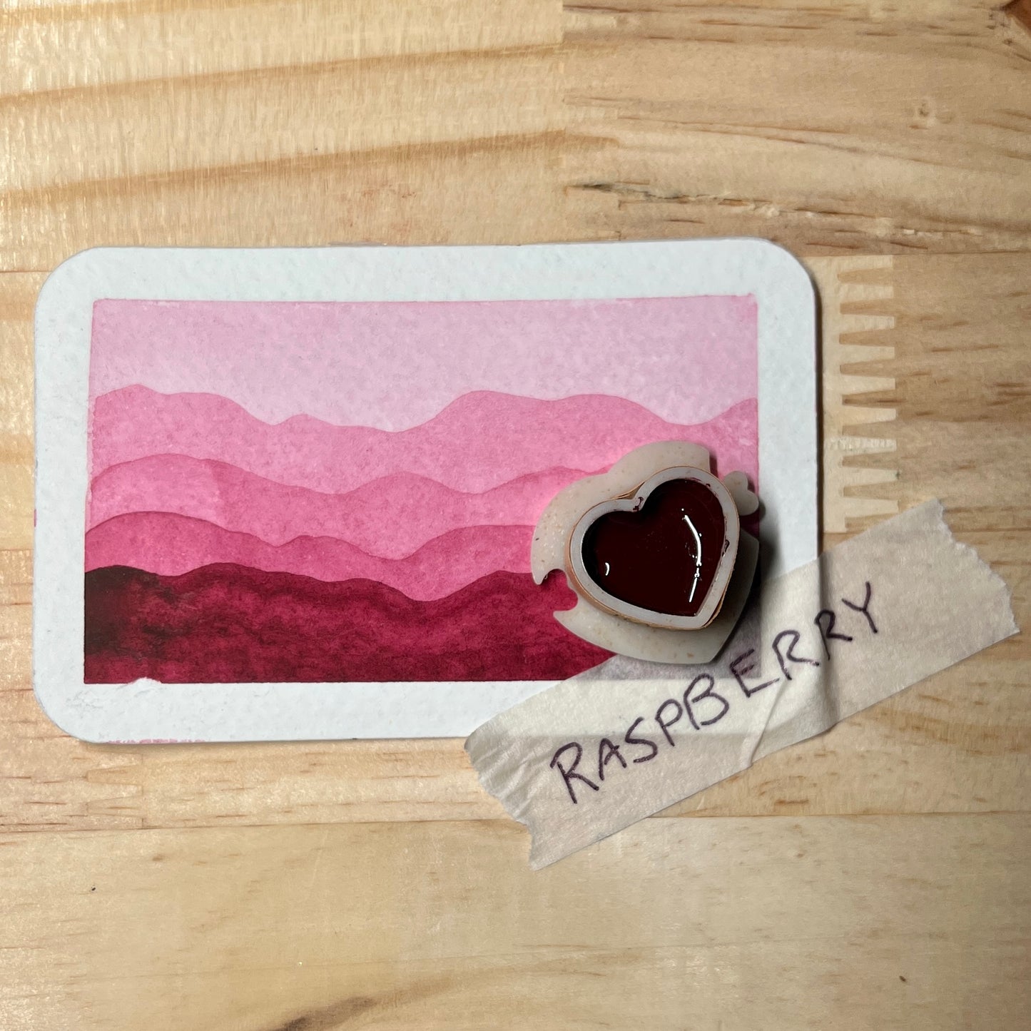Heart-shaped watercolor pan with  raspberry pink color on it  ontop of a gradient swatch card with label 'Raspberry' on a wooden surface