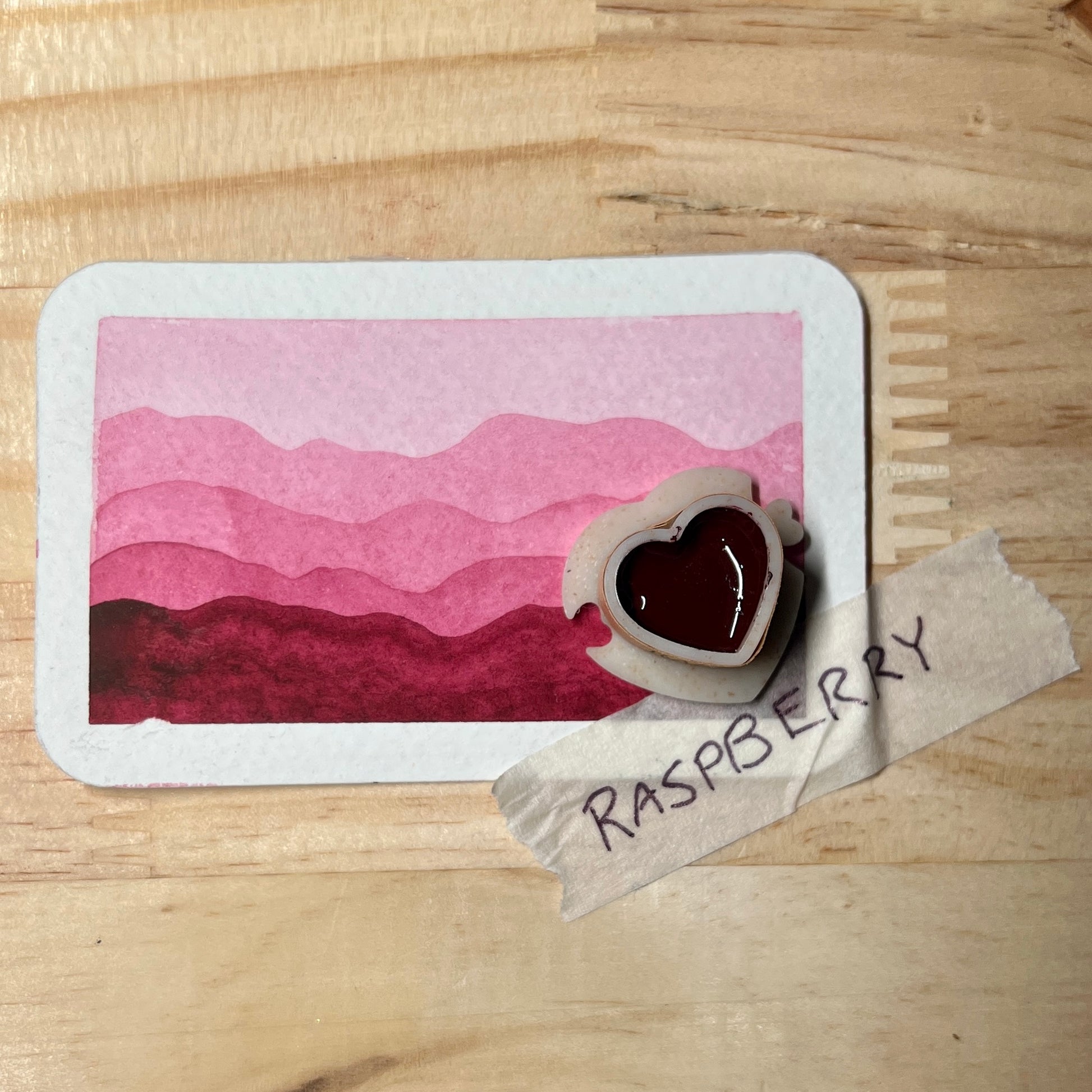 Heart-shaped watercolor pan with  raspberry pink color on it  ontop of a gradient swatch card with label 'Raspberry' on a wooden surface